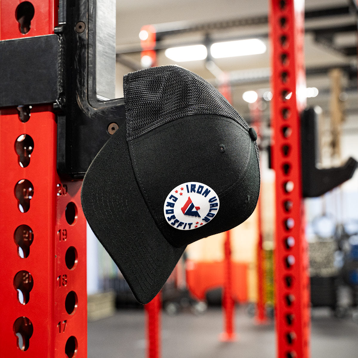 Black cap with a logo hanging on a red weightlifting rack in a gym.
