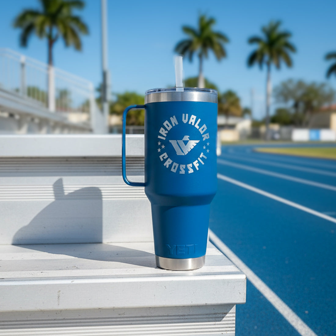 Blue insulated mug with handle and straw lid on a white ledge with palm trees and a track in the background